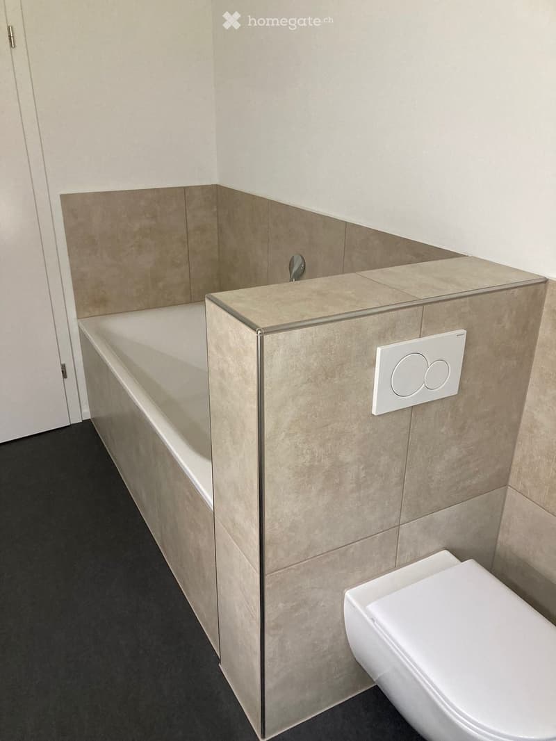 Modern bathroom with a bathtub, toilet, and a sleek faucet. The walls are tiled in a light beige color, and the floor is dark.