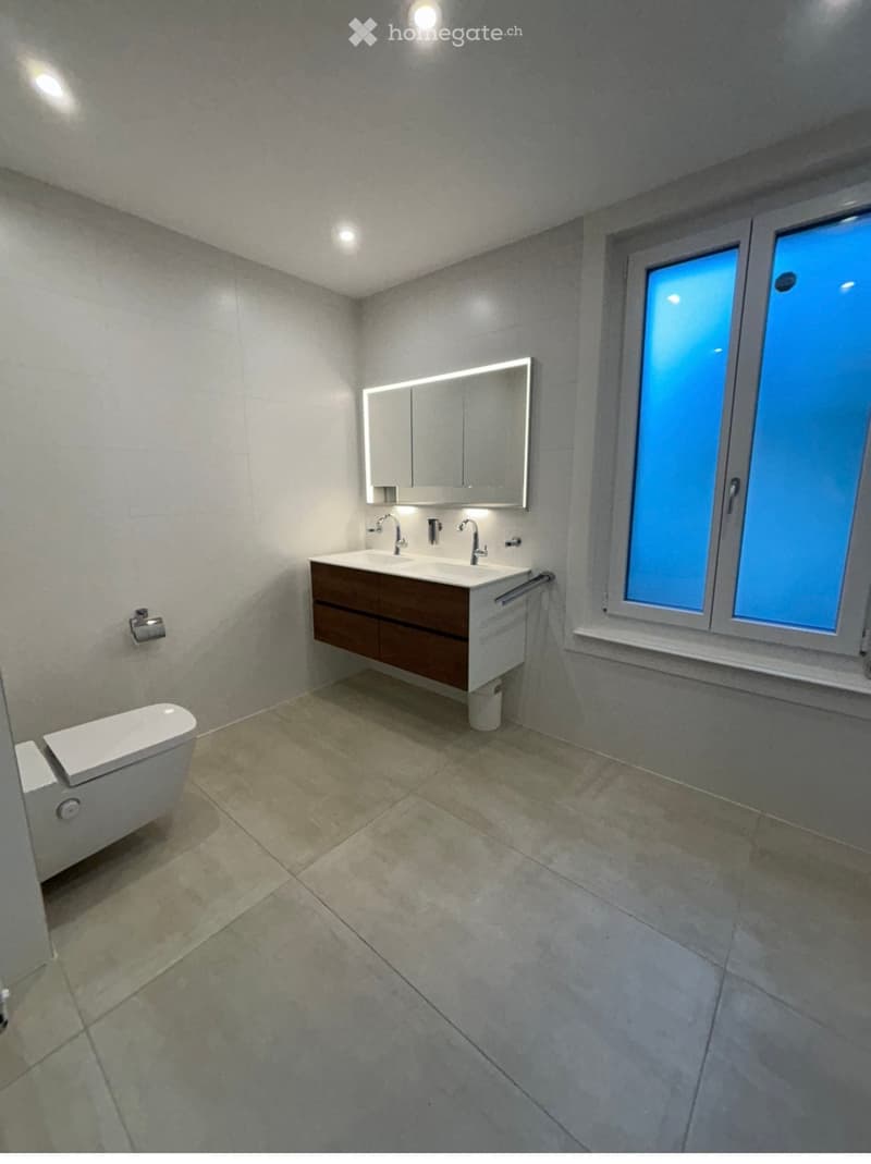 Modern bathroom with a double sink, toilet, large mirror, and built-in cabinetry. The space features tiled floors and walls with recessed lighting.