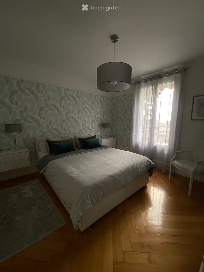 A well-lit bedroom with a large bed, nightstands, a chair, and a hanging lamp. The room features wooden flooring and patterned wallpaper on one wall.