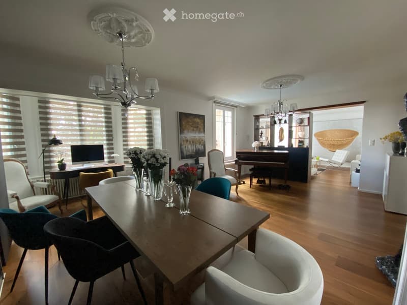 A spacious, well-lit room with wooden flooring, large windows, and modern decor. Features include a long wooden dining table with multiple chairs, a piano, a computer setup, and decorative vases with flowers.