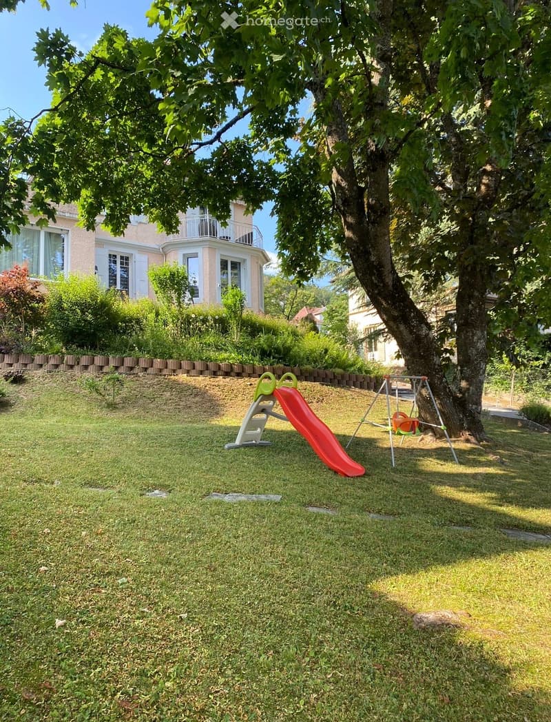 A well-maintained garden with a slide, swing, and a large tree providing shade. The lawn is surrounded by a low brick wall and a house with a balcony is visible in the background.