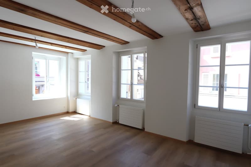 Empty room with wooden flooring, white walls, exposed wooden beams on the ceiling, and multiple windows with white frames. Radiators are visible beneath the windows.