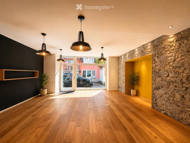 Empty room with wooden flooring, large windows, and modern pendant lights. Features a stone wall and a yellow accent wall.