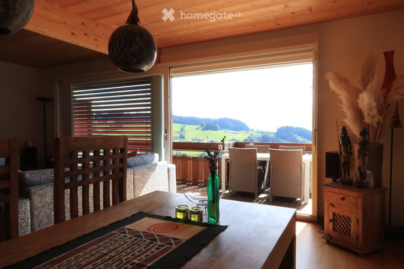 Furnished Room. Bright living room with wooden ceiling, large windows, and a view of the countryside. Features a sofa, dining table, chairs, decorative vase, candles, and statues.