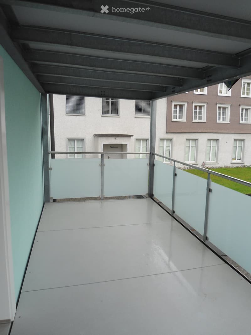 Modern balcony with white tiled floor, glass railings, and a view of neighboring buildings and greenery.