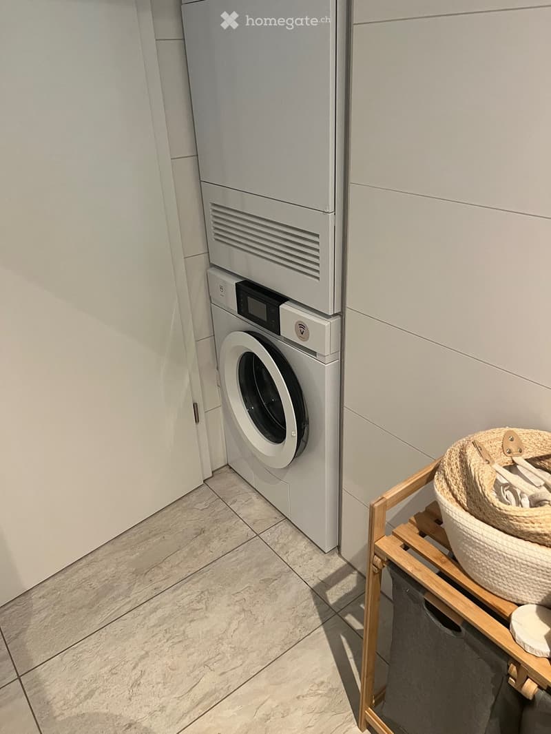 A modern bathroom with a white washing machine, a drying rack, and a wicker basket on a wooden shelf.