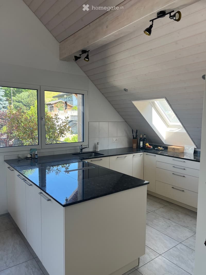 Modern kitchen with white cabinets, black countertops, and a large window. Features include a sink, faucet, wine bottles, fruit basket, and cutting board on the counter.