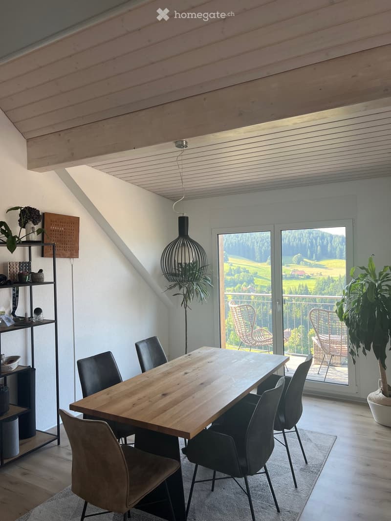 Bright dining area with wooden table, black chairs, modern pendant light, and large window offering scenic view.