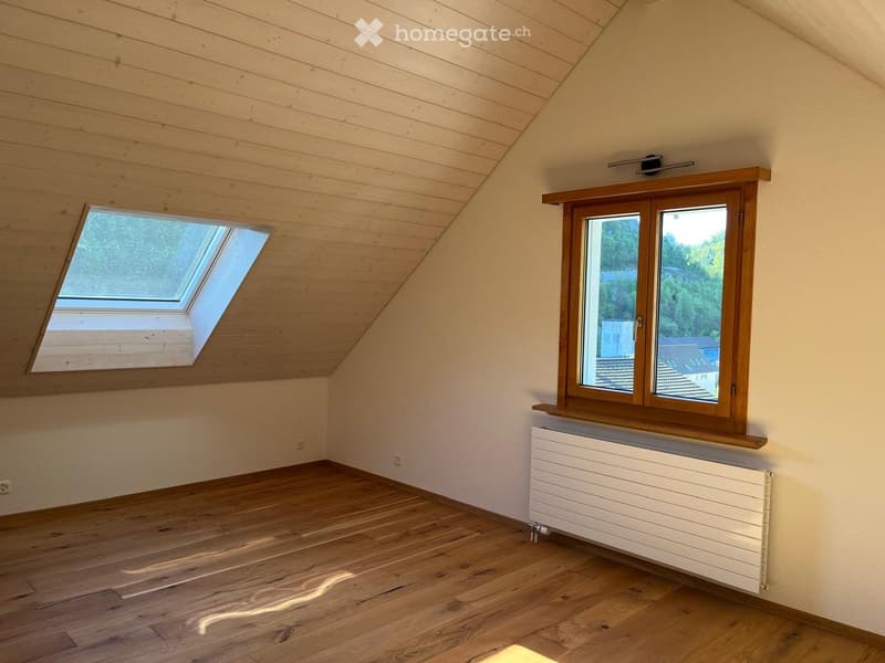 Empty attic room with wooden floors, slanted wooden ceiling, skylight window, and double-hung window with radiator below.