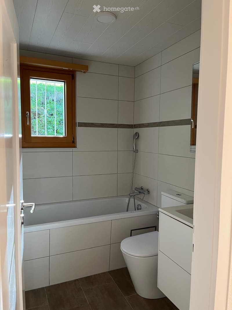 Modern bathroom with a bathtub, toilet, sink, and showerhead. Grey tiled walls and wooden ceiling.