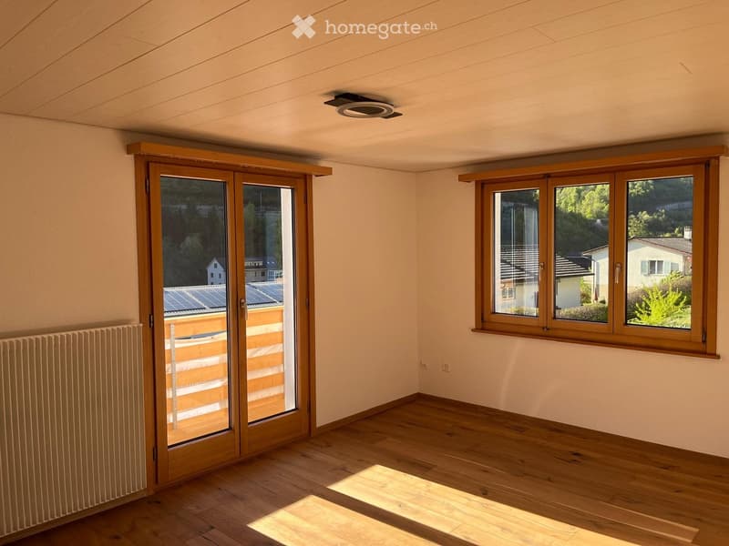 Empty room with wooden flooring, large windows, and a sliding glass door leading to a balcony. The room has light-colored walls and a visible radiator on one side.