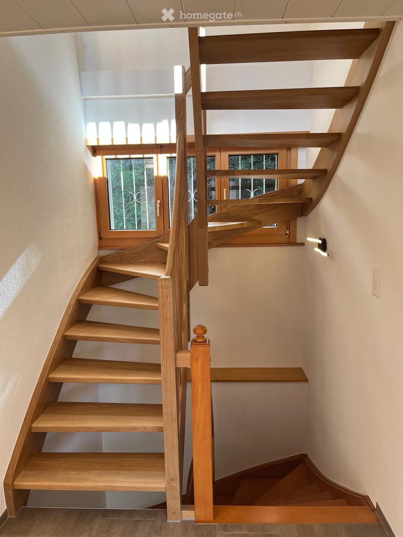 Spiral staircase with wooden steps and handrail, illuminated by natural light from a window, leading to an upper level.