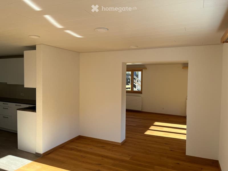 Empty room with wooden flooring, white walls, and a small kitchen area visible in the background. The space is well-lit with natural light coming through a window.