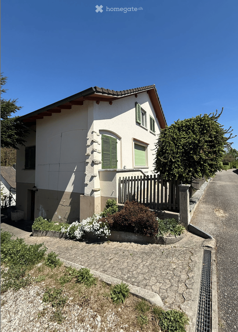 Two-story house with white exterior, green shutters, and a small garden area with shrubs and flowers.