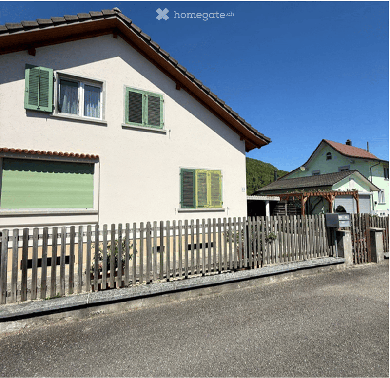 Single-story house with white walls, green shutters, and a wooden fence in front. The roof is sloped with red tiles.