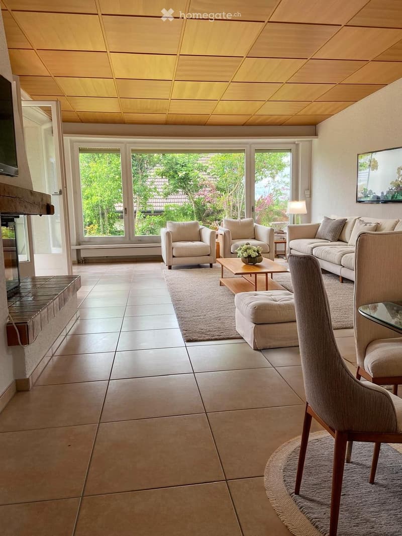 Bright living room with large windows and a view of greenery, featuring a beige sofa, armchairs, coffee table, ottoman, and a fireplace.