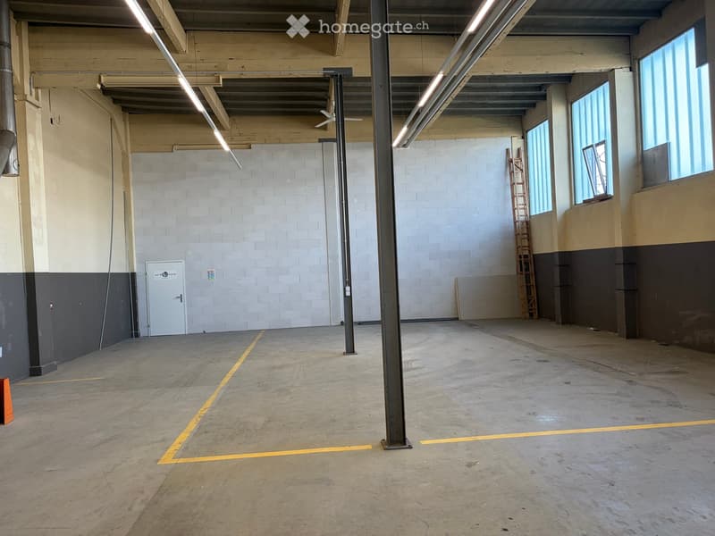 Empty industrial-style room with concrete floor, exposed brick walls, large windows, and metal support columns.