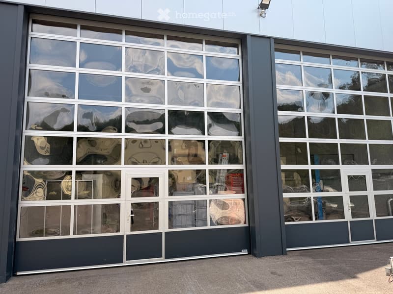 Modern building with large glass garage doors, reflecting the sky and surrounding structures. The entrance features multiple panels and a clean, contemporary design.