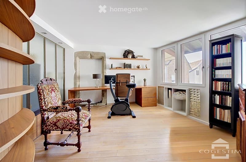 A well-lit room with wooden flooring, large windows, a patterned armchair, a wooden desk with a laptop, a lamp, a bookcase, and an exercise bike.