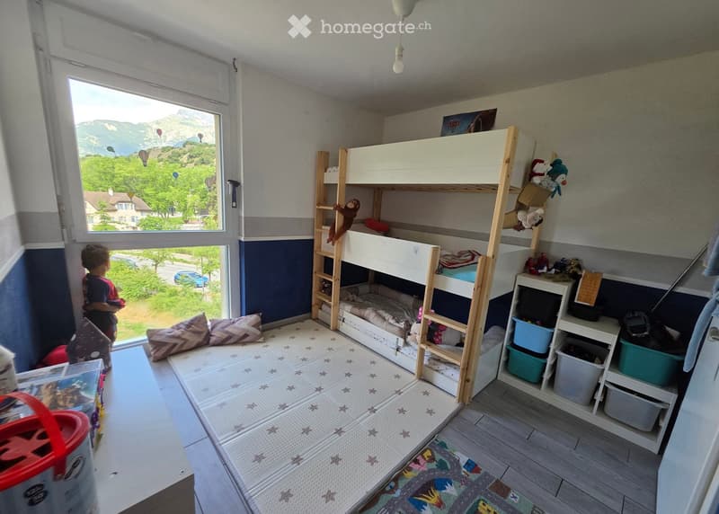 A well-lit bedroom with a bunk bed, shelves, toys, and a view of the mountains through a large window.