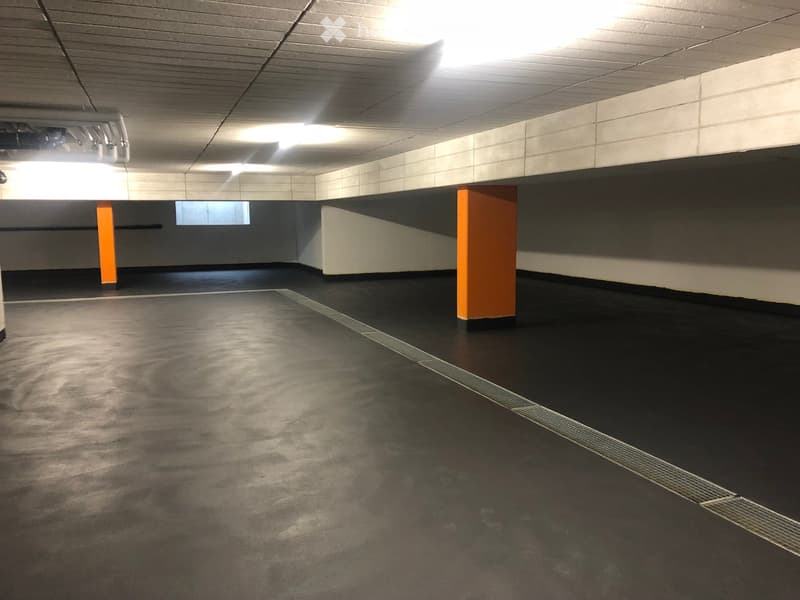 Underground parking area with black flooring, white tiled walls, and orange pillars. The space is empty with overhead lighting and a small window at the far end.