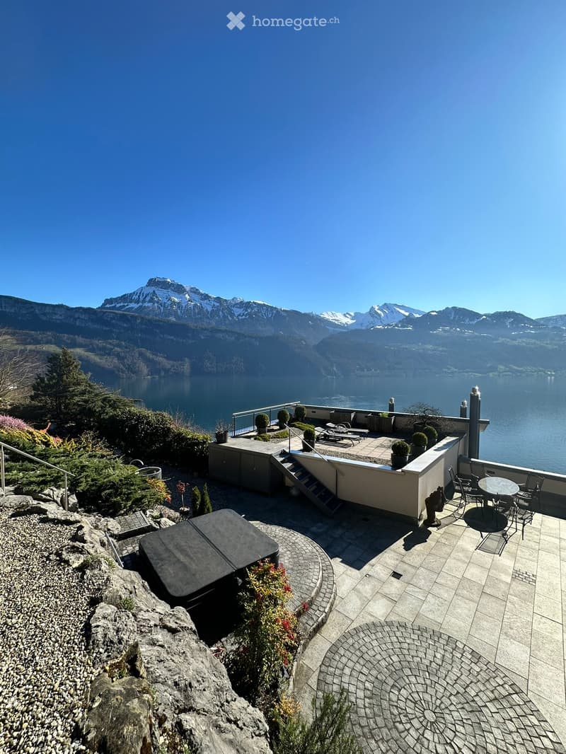 Outdoor terrace with a panoramic mountain and lake view, featuring a table, chairs, sun loungers, potted plants, and a hot tub.