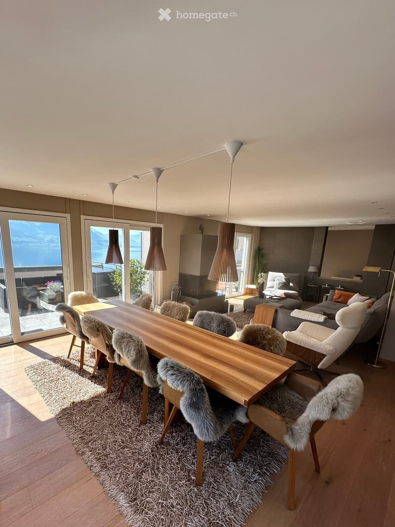 Modern dining area with a large wooden dining table, fur-covered chairs, a plush rug, and a cozy living area with a sofa and armchair, overlooking a scenic lake view.