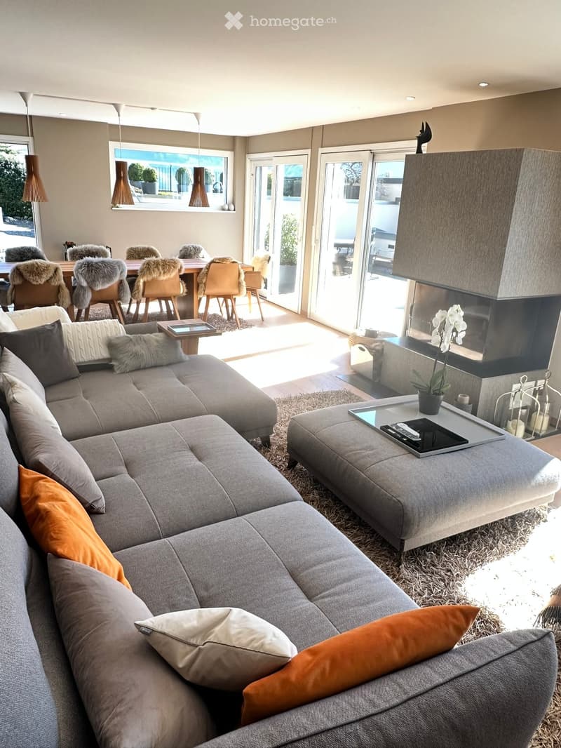 Modern living room with a grey sectional sofa, coffee table, ottoman, pendant lights, dining area with chairs, fireplace, and large windows offering a view.