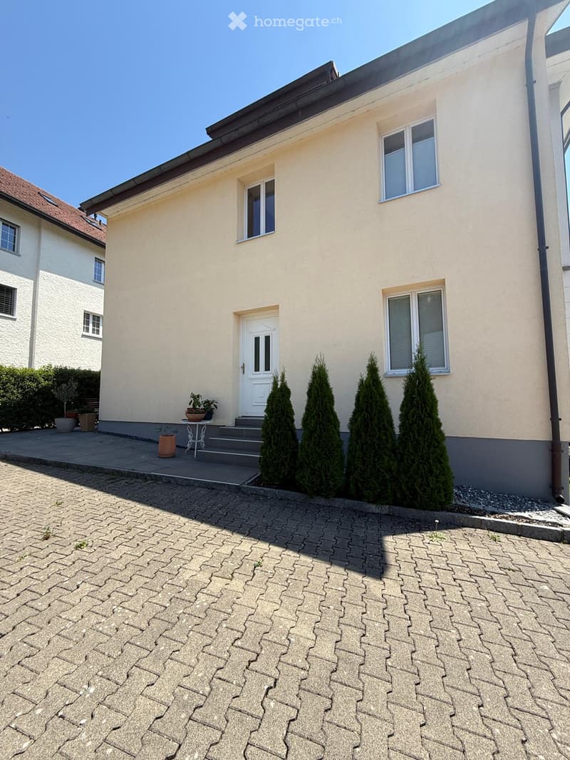 Two-story house with beige exterior, white door, multiple windows, and small garden area with potted plants and a table.