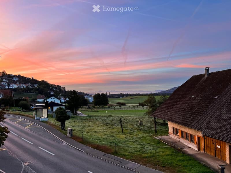 A rural house with a tiled roof and wooden shutters, situated on a grassy field near a road, with a scenic sunset view of hills and scattered houses in the background.
