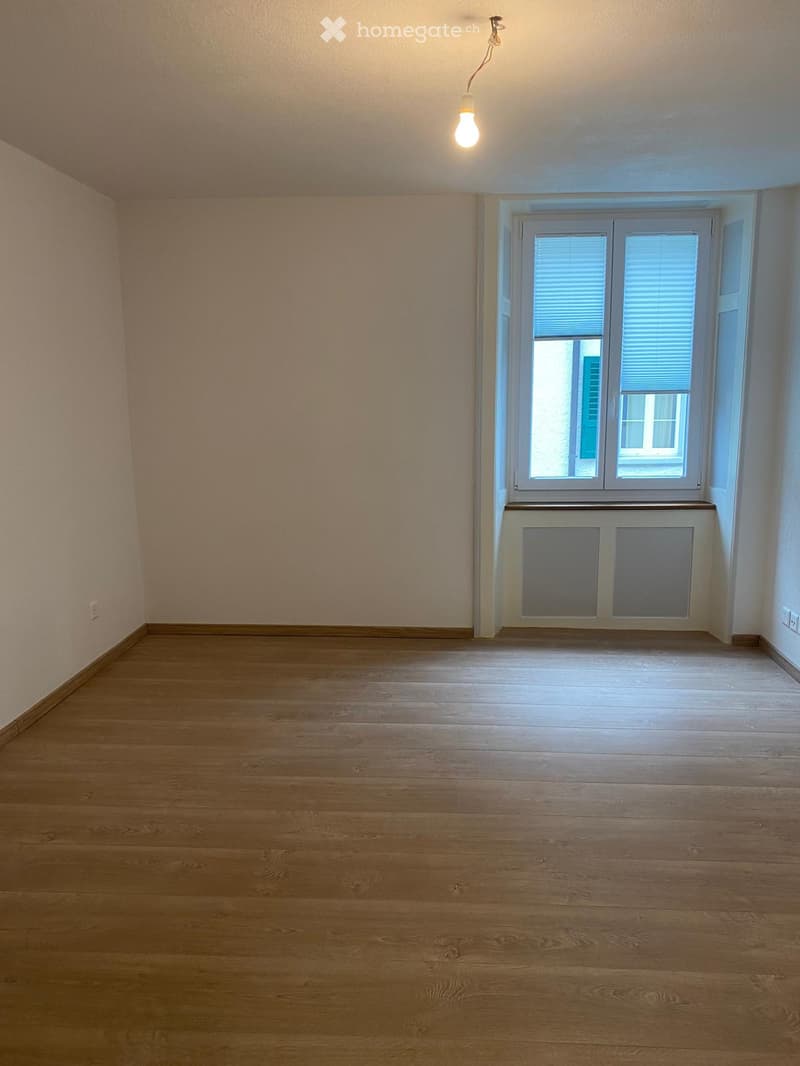 Empty room with wooden flooring, light-colored walls, and a window with blinds. A radiator is visible below the window, and a single light fixture hangs from the ceiling.