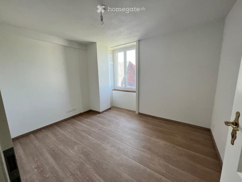 Empty room with light wooden flooring, white walls, and a window with a radiator below it. The room has a simple, clean design with no furniture or decorations.