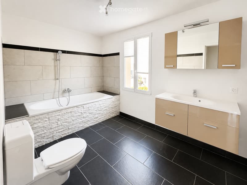 Modern bathroom with a bathtub, toilet, sink, mirror, and cabinet. Features black tiled flooring and beige tiled walls with a black accent border.