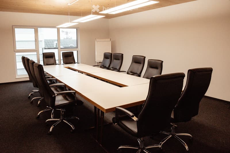 Conference room with a long rectangular table surrounded by black office chairs, large windows, and a whiteboard on the wall.