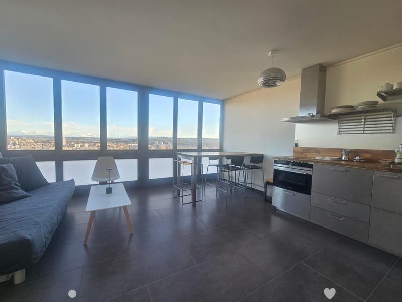 Modern open-plan living and dining area with a sofa, small table, dining table with chairs, and a kitchen area with an oven and cabinets. Large windows offer a scenic view.
