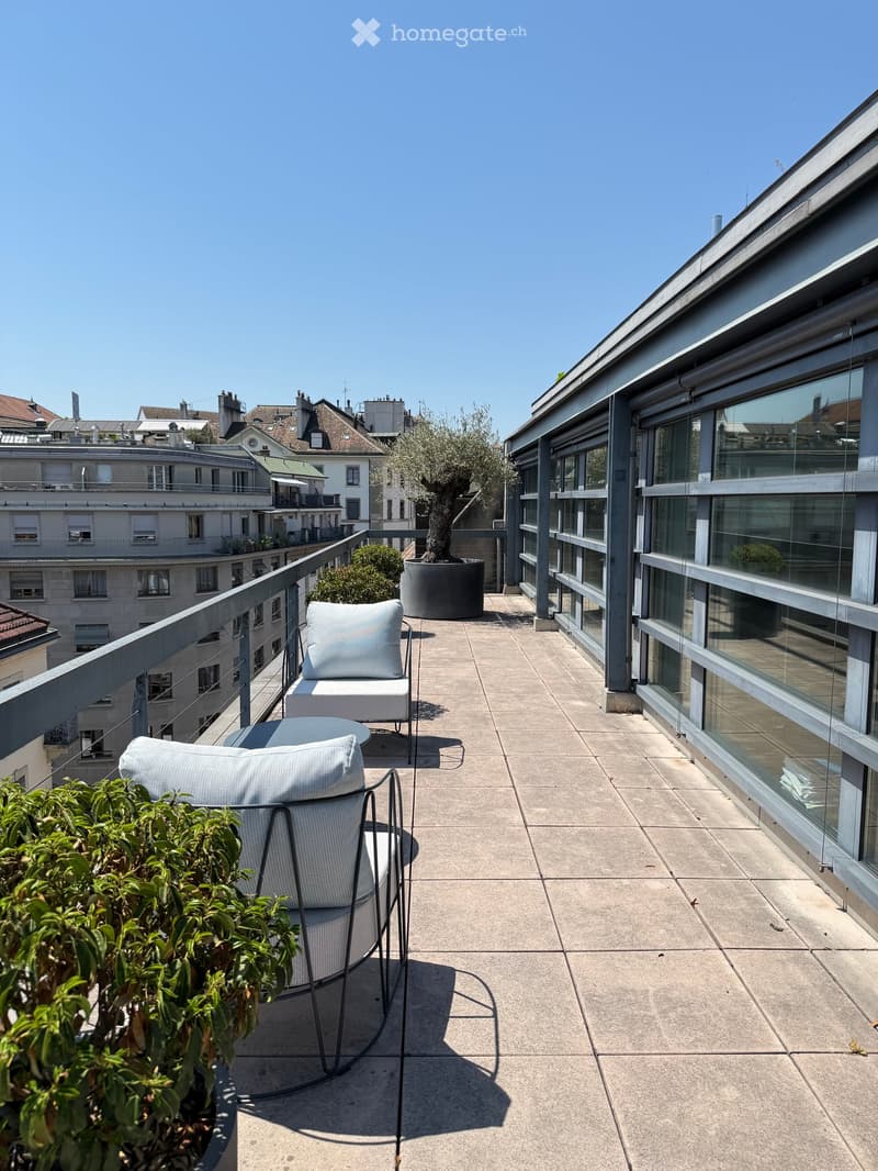 Modern rooftop terrace with a concrete floor, glass railing, and a view of the cityscape. Features a white chair, small table, and potted plants.