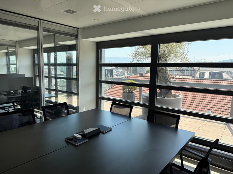 Modern office space with large windows, black conference table, chairs, laptop, and office equipment.
