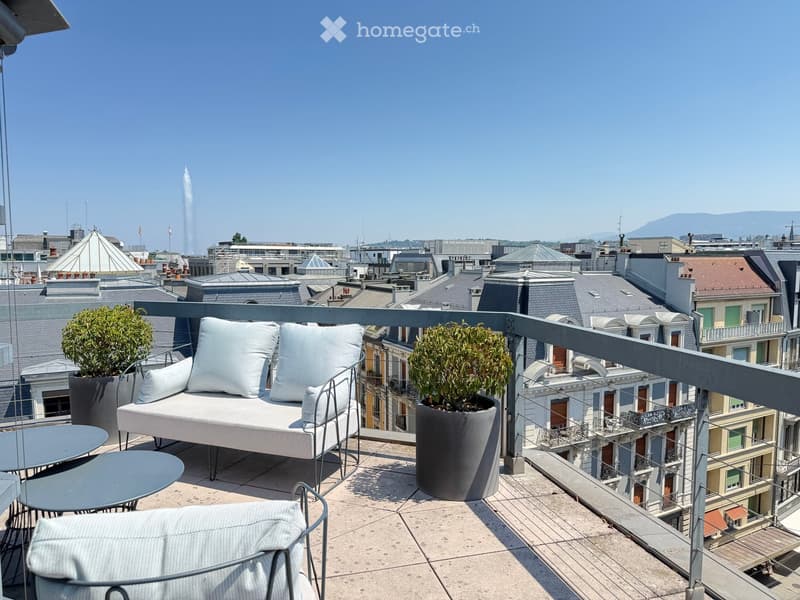 A modern rooftop terrace with a white sofa, two side tables, a chair, and potted plants, overlooking a cityscape with rooftops and a fountain.