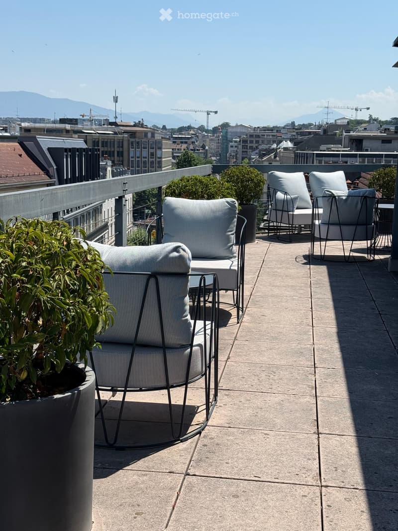 Modern balcony with grey chairs, small table, and potted plants, overlooking a cityscape with mountains in the background.