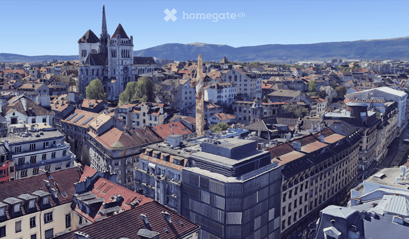 A panoramic view of a historic cityscape with numerous buildings, red-tiled roofs, and a prominent church tower in the background, surrounded by mountains under a clear blue sky.