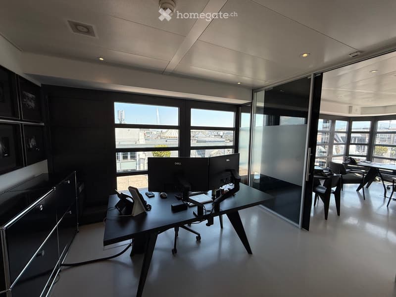 Modern office space with large windows, two desks, multiple monitors, chairs, and a screen divider.
