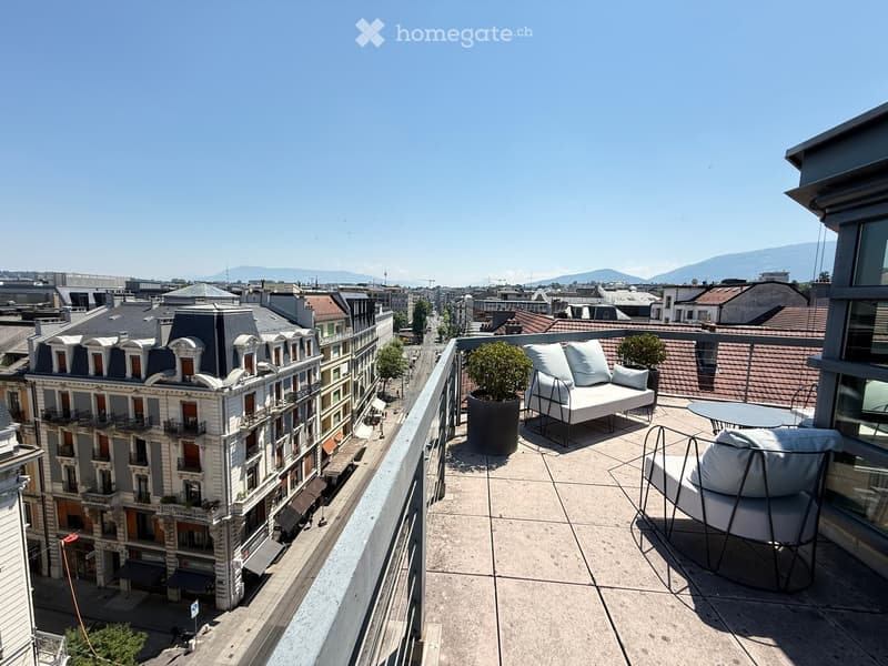 Balcony with a modern sofa, chairs, a small table, and potted plants, overlooking a cityscape with historic buildings and distant mountains.