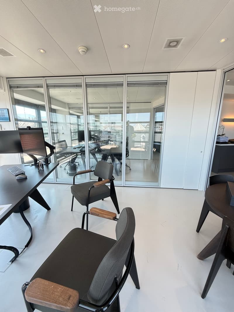 Modern office space with white walls, large glass windows, and contemporary furniture including chairs, tables, and a computer setup.