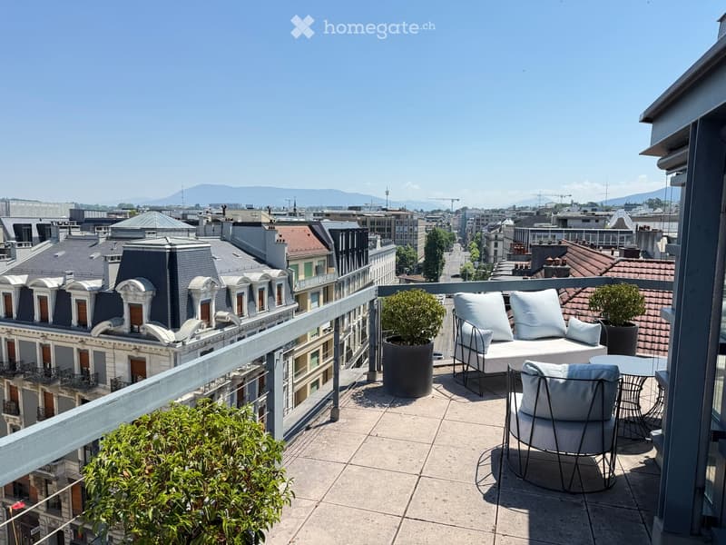 Balcony with a modern seating area, including a sofa, two chairs, a small table, and potted plants, overlooking a cityscape with rooftops and distant mountains.