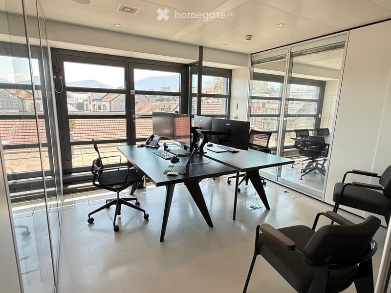 Modern office space with large windows, a black desk, multiple chairs, and computer equipment.