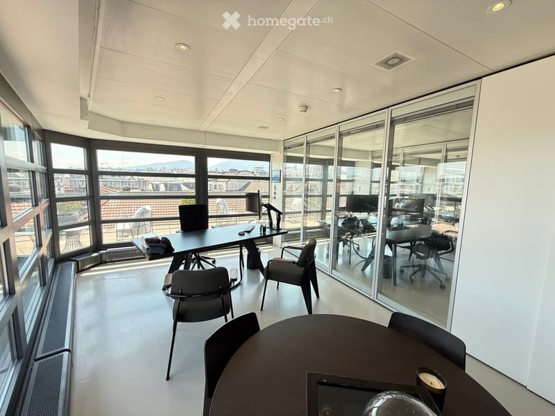 Modern office space with large windows offering a city view, featuring desks, chairs, and a round meeting table.