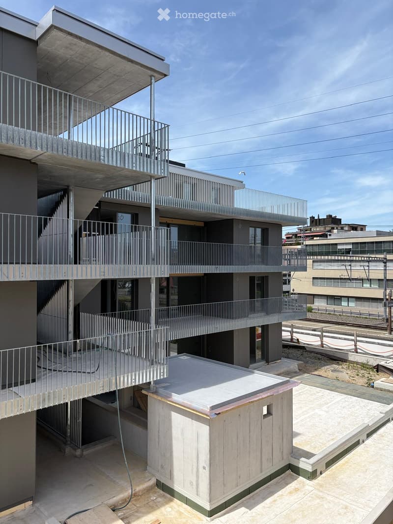 Modern multi-story building with balconies, metal railings, and a rooftop area featuring a small wooden structure and concrete surfaces.