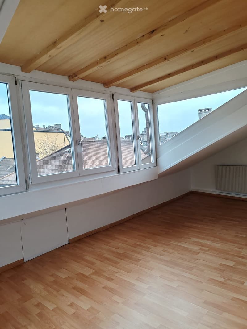 Empty attic space with wooden flooring, large windows offering a view of rooftops, and exposed wooden beams on the ceiling.