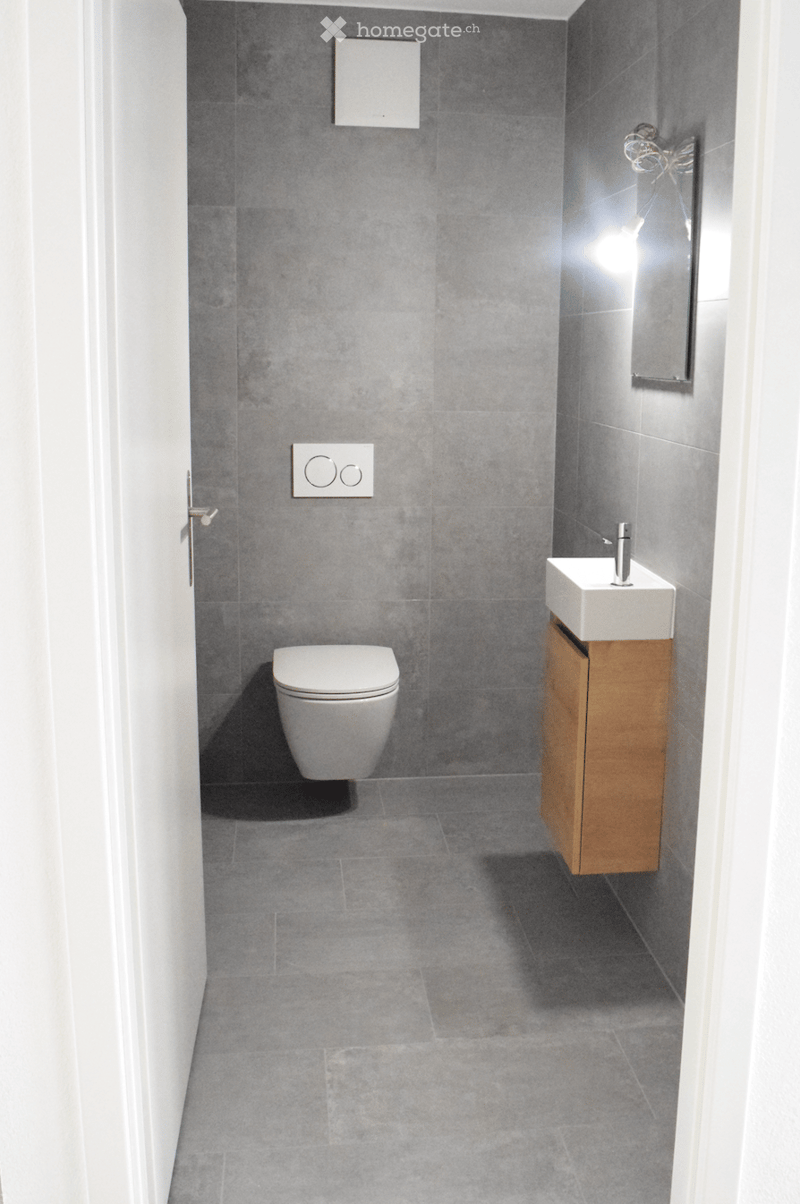 Modern bathroom with grey tiled walls and floor, white toilet, white sink with wooden cabinet, and a mirror above the sink.