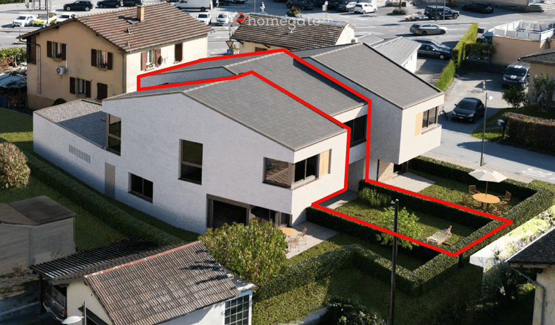 Aerial view of a modern two-story house with a grey roof, large windows, and a garage. The house is surrounded by a well-maintained garden with outdoor seating and a parking area.